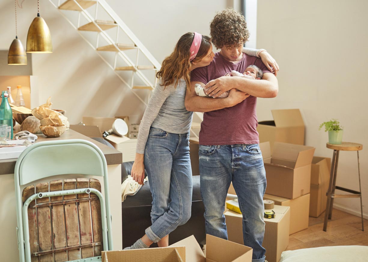 A young couple are holding an infant in a home they recently purchased with a new home mortgage