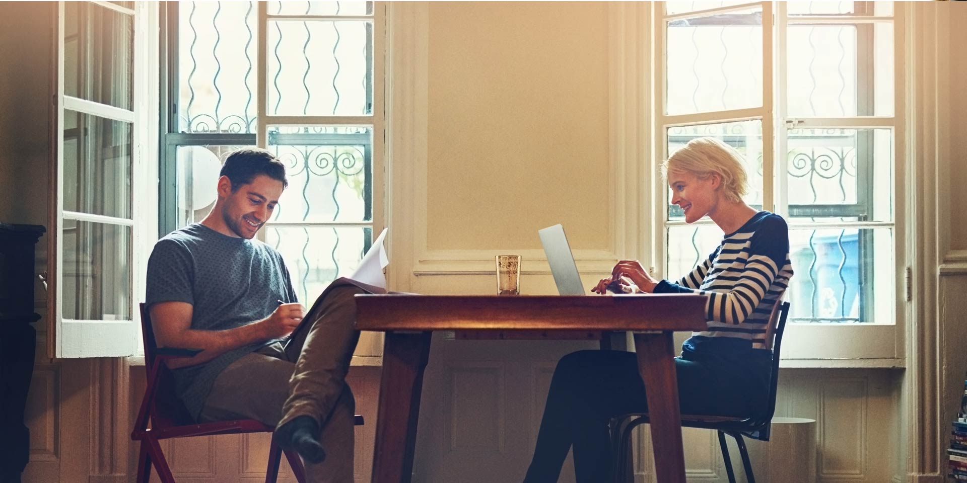 A younger couple sitting at a kitchen table are working on laptops and discussing CD rates